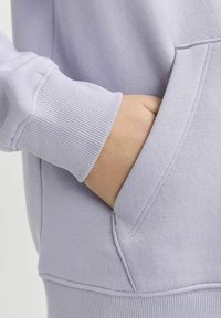 Hoodie di un leggero colore viola con polsini a costine, tasca frontale e finitura testurizzata. Presenta cuciture visibili lungo le cuciture.