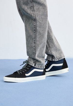 Baskets en toile noire et marine avec des accents blancs, des lacets texturés et un bout arrondi. Portées avec un jean gris déchiré, posées sur un fond bleu.