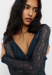 Frau mit langen dunklen Haaren, die ein tief ausgeschnittenes türkisfarbenes Spitzenoberteil trägt und die Arme vor der Brust verschränkt hat, während sie leicht zur Seite blickt, vor einem weißen Hintergrund.