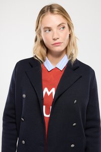 Veste bleu marine à double boutonnage avec des boutons argentés, en tissu texturé, portée sur un sweat-shirt rouge avec des lettrages blancs, superposée à une chemise à col rayée bleue.