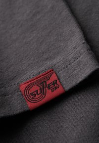 Tessuto di maglietta in cotone grigio con un'etichetta rossa visibile che presenta "SUPER DRY" e un design di ali, enfatizzando la texture e i dettagli delle cuciture.