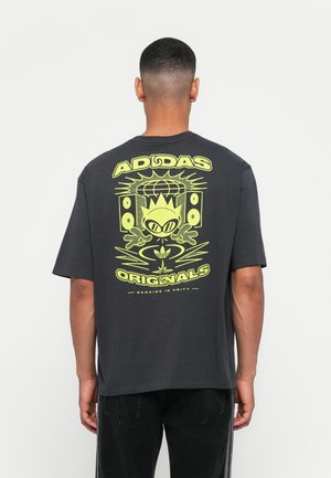Egy férfi hátulról látható, fekete Adidas Originals pólóban, neon sárga grafikával, amelyen egy mesefigura, hangszórók és egy gömb látható.