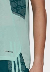 Detail zblízka na osobu v světle modrém sportovním topu z síťoviny s nápisem "PRIMEBLUE" a tmavě modrých šortkách s vzorem a světle modrými pruhy.