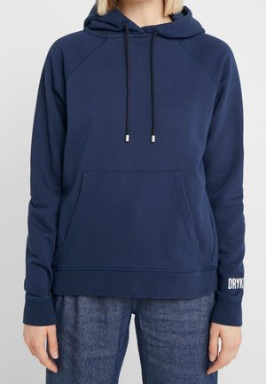 Personne portant un sweat à capuche bleu marine avec poche frontale et cordons noirs, assorti à un jean bleu foncé. Logo Drykorn sur le bas de la manche gauche.