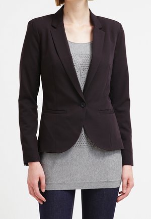 Žena v černém přiléhavém saku přes šedý svetřík s cvočky a tmavě modrých džínách stojí před jednobarevným bílým pozadím.