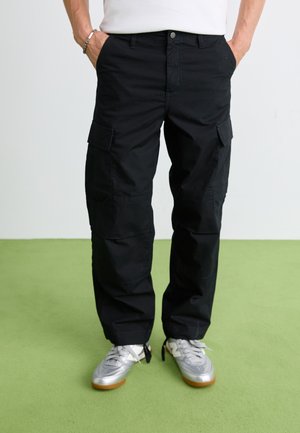 Hombre con pantalones cargo negros y zapatillas plateadas de pie sobre un suelo verde con las manos en los bolsillos, contra un fondo blanco.