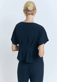 Una persona con capelli biondo chiaro raccolti in un chignon indossa una maglietta blu navy a maniche corte con un design peplum, girata di spalle rispetto alla macchina fotografica.