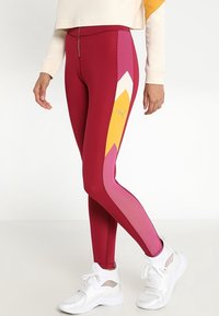 Leggings atletici rossi con motivi geometrici gialli e rosa, dettagli in rete e un tessuto liscio e elasticizzato. Abbinati a sneakers bianche.