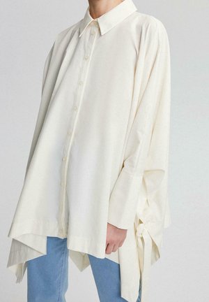Oversized off-white skjorte med asymmetrisk kant og bindedetalje i siden, båret med lyseblå jeans.