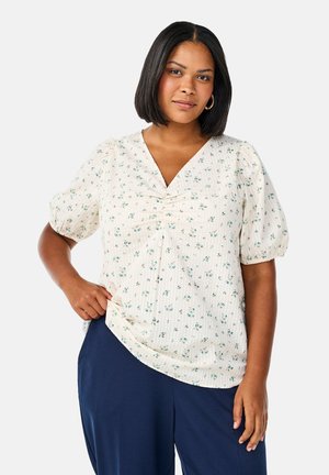Blouse blanche à fleurs avec manches bouffantes, décolleté en V et détails froncés. Le tissu présente un motif texturé et de petites fleurs vertes.