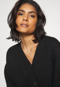 Svart stickad cardigan med V-hals, med stora knappar och en strukturerad tyg. Silverhalsband med en liten berlock kompletterar looken.