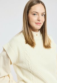 Femme avec des cheveux lisses châtain clair portant un pull sans manches en tricot à torsades crème par-dessus une blouse légère, légèrement de profil vers la droite, sur un fond neutre.