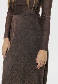 Eine Frau in einem langen, glitzernden Kleid in Dunkelbraun steht mit entspannten Händen an ihrer Seite vor einem schlichten Hintergrund.