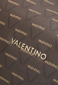 Barna textúrájú bőrtáska, amelyen a "Valentino" logók dombornyomottak, és egy kiemelkedő arany tónusú logó díszíti.