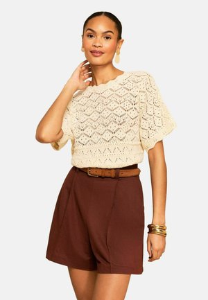 Žena má na sebe krémový háčkovaný top s krátkym rukávom, hnedé krátke nohavice s vysokým pásom a opaskom, zlaté náušnice a náramky zložené do vrstiev.