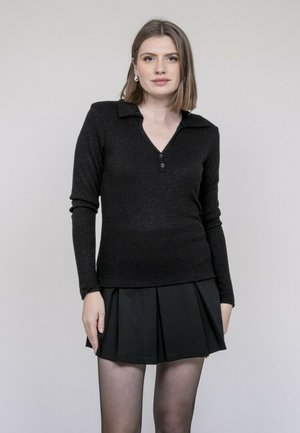 Jeune femme aux cheveux mi-longs portant un haut en maille à manches longues noir et une jupe plissée noire, debout devant un fond uni.