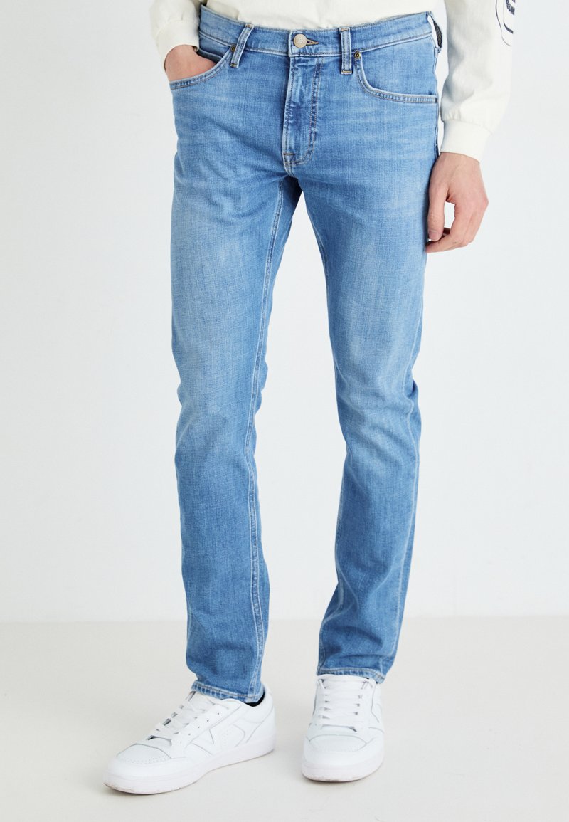 Világoskék farmernadrág, slim fit fazonnal, öt zsebbel és látható varrással. Fehér sportcipőkkel viselve, amelyek texturált felülettel rendelkeznek.