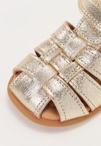 Metallic guld sandal med krydsede stropper, åben tå og en struktureret overflade. Sålen er tanfarvet og giver kontrast til den metalliske overdel.