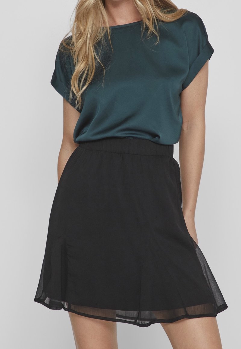 Femme portant un chemisier court à manches courtes de couleur bleu sarcelle foncé, rentré dans une jupe noire transparente arrivant au genou, sur fond gris.