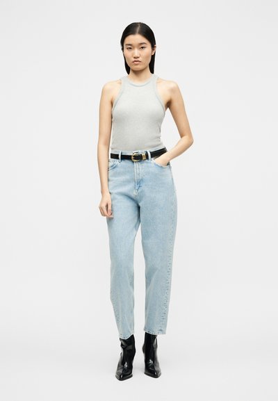 Νεαρή γυναίκα όρθια με το ένα χέρι στην τσέπη, φοράει γκρι αμάνικο τοπ, ανοιχτόχρωμο τζιν ψηλόμεσο, μαύρη ζώνη και μαύρα μποτάκια.