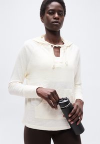 Άτομο που φοράει μπεζ φούτερ με κουκούλα και σκούρο παντελόνι, κρατώντας ένα μαύρο μονωμένο παγούρι νερού με το ένα χέρι στο καπάκι και το άλλο στο σώμα.