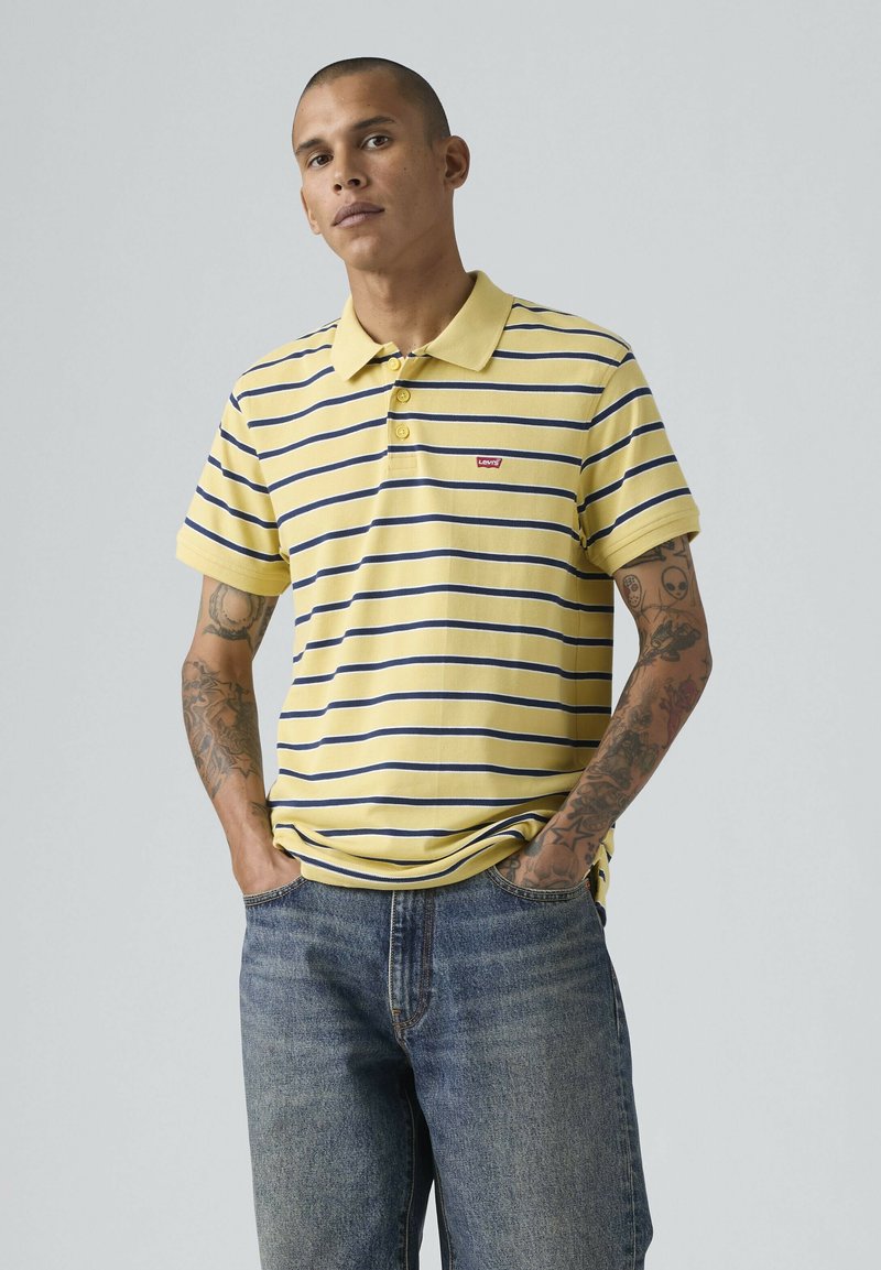 Homme tatoué portant un polo rayé jaune et marine et un jean bleu, debout avec les mains dans les poches devant un fond uni.