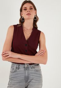 Bordová bezrukávová blúzka s výstrihom do V, s detailom gombíka a textúrovanou tkaninou. Skombinovaná so svetlo šedými džínsami s vysokým pásom.