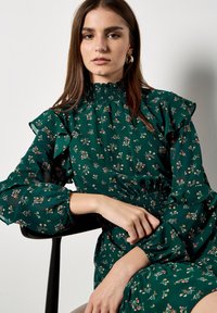 Zielona sukienka w kwiaty z falbaniastymi rękawami, wysokim marszczonym dekoltem i dekoracyjnymi wzorami kwiatowymi. Gładka tekstura materiału z marszczonym pasem.