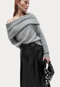 Oversized šedý pletený svetr s odkrytou šíjí, kombinovaný s černou koženou sukní; modelka drží černou kabelku zdobenou stříbrnými hroty.
