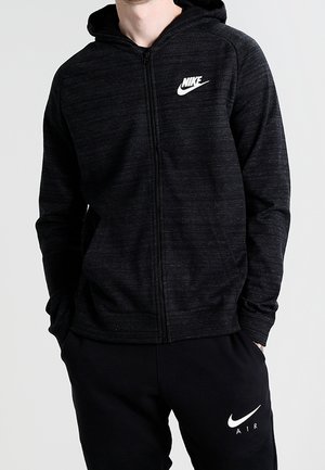 Mężczyzna w czarnej bluzie Nike zapinanej na zamek i czarnych spodniach dresowych Nike Air, stojący z rękami w kieszeniach na jednolitym tle.