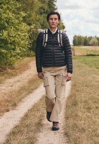 Giacca a vento nera, pantaloni cargo beige e scarponi da trekking marroni. Design robusto, tasche visibili sui pantaloni, che cammina su un sentiero erboso e sterrato.