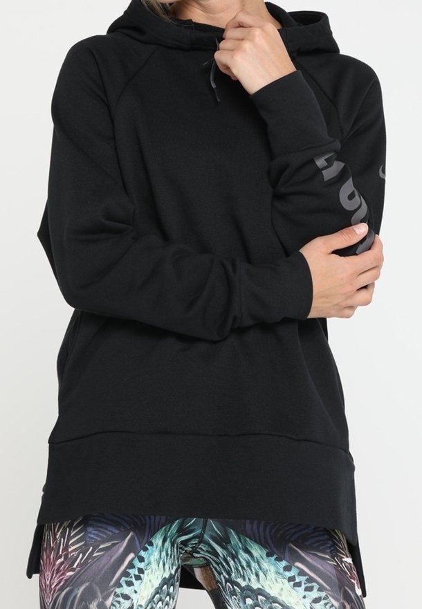 Personne portant un sweat à capuche noir avec un texte partiel sur le bras, tenant un bras, et un legging à motif abstrait bleu et vert.