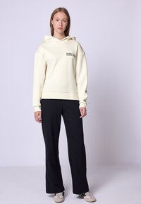 Felpa crema con logo frontale, abbinata a pantaloni neri a gamba larga. Il tessuto è morbido con una texture opaca; scarpe sportive dallo stile casual completano il look.