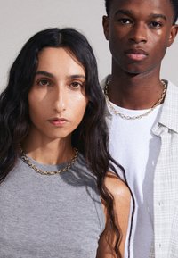Guld kædetråde båret af to personer; en i en grå tanktop, den anden i en hvid skjorte og en lys rutet jakke.