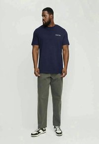 Uomo che indossa una maglietta Club1990 blu navy, pantaloni grigi e sneakers bianco e nero, in piedi e guarda a sinistra.