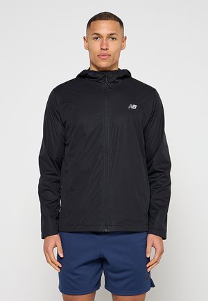 Joven con chaqueta negra con capucha y cremallera con el logo de New Balance y pantalones cortos deportivos azul marino, de pie mirando hacia adelante.