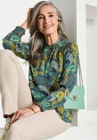 Langsleeve, gemusterde blouse in blauw, groen en goud met een gerimpelde hals, gecombineerd met beige broeken en een mintgroene handtas.
