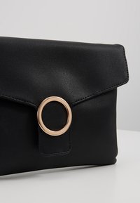 Svart läderkuvert med lockstängning och en accent av guldmetallring. Slät textur med kontrasterande sömnadsdetaljer.