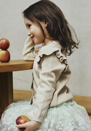 Jeune fille en jupe fleurie et pull beige assise à une table en bois, tenant une pomme et regardant deux pommes empilées sur la table.