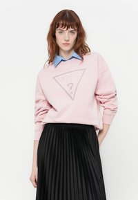 Mujer con cabello rojo vistiendo un sudadera rosa de Guess sobre una camisa de cuello azul y una falda plisada negra, de pie frente a un fondo blanco.