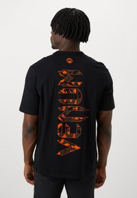 Sort t-shirt med stor orange camouflagetekst "MEDIUM" lodret på ryggen, med et lille logo ved halsen. Korte ærmer.