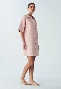 Krótka różowa sukienka zapinana na guziki z kołnierzykiem, krótkimi rękawami, dwoma przednimi kieszeniami i luźnym krojem, wykonana z lekkiej tkaniny.