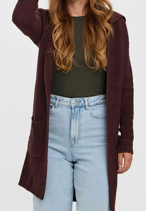 Femme portant un haut côtelé vert foncé, un jean taille haute bleu clair et un long cardigan texturé violet foncé avec des poches.