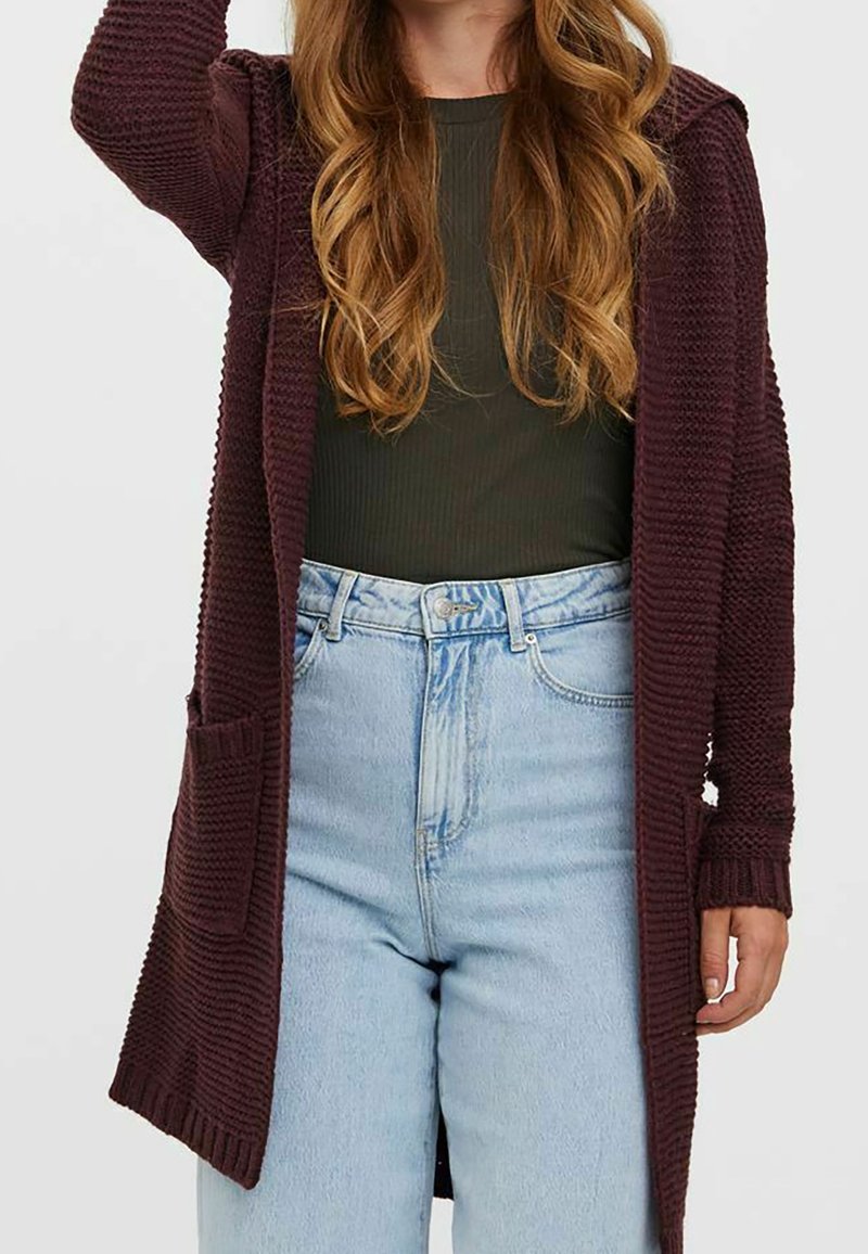 Femme portant un haut côtelé vert foncé, un jean taille haute bleu clair et un long cardigan texturé violet foncé avec des poches.