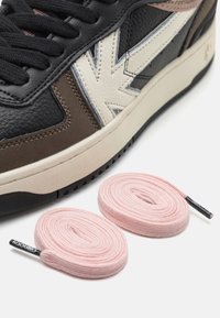 Černé a hnědé kožené tenisky s krémovým akcentem a růžovými tkaničkami na bílém pozadí. Mají texturované materiály a nízký profil.