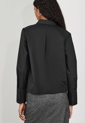 Vrouw met lang krullend haar die een zwarte blouse met lange mouwen en een grijze gestructureerde rok draagt, gezien van achteren tegen een effen achtergrond.