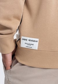 Beżowa bluza z prążkowanymi mankietami i dołem, z etykietą z napisem "OMBRE MENSWEAR" w czarnym kolorze na białym tle.