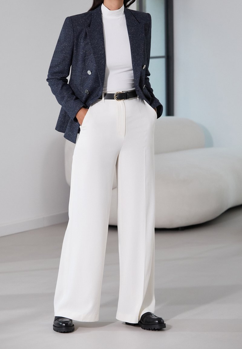 Personne portant un pantalon blanc à jambes larges, une ceinture noire, un haut blanc à col montant, un blazer sombre croisé et des chaussures noires, debout à l'intérieur.