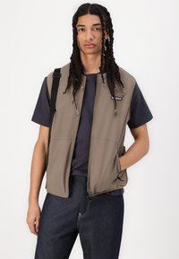 Brown softshell vest with a zip front, side pockets, and a logo detail. Worn over a dark t-shirt and blue jeans, featuring a relaxed fit.