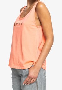 Top de tank peach hecho de una tela suave, con un corte relajado, escote redondo y un logo impreso en un tono oscuro, combinado con pantalones grises.